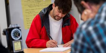 Manin white shirt and red jacket writing at table.