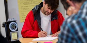 Man in red jacket writes in notebook at table.